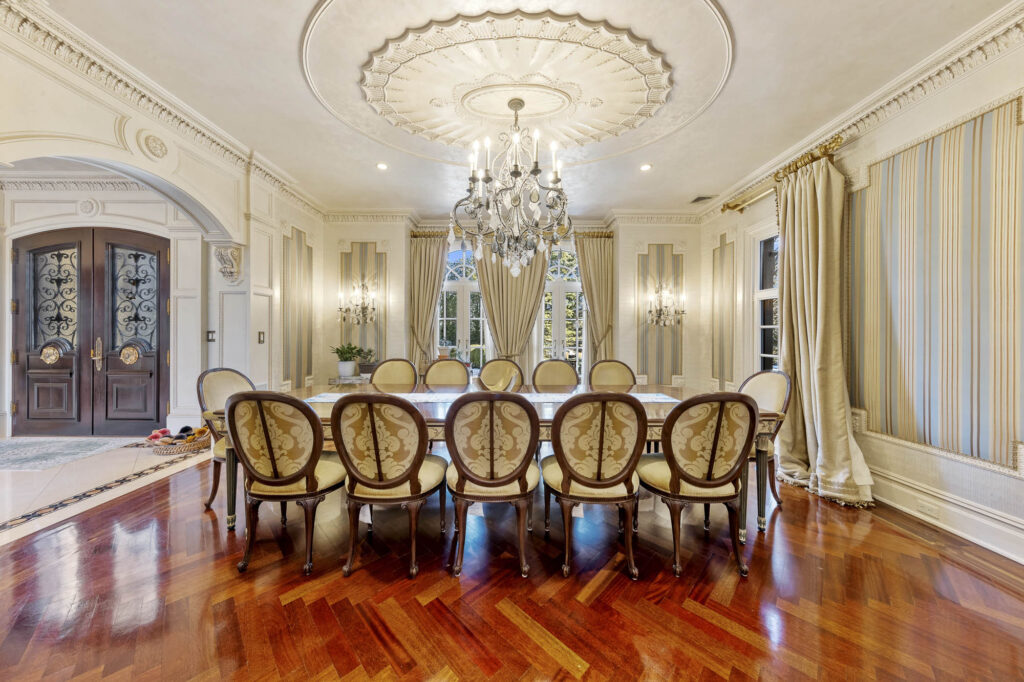 Elegant dining room with a long table surrounded by ornate chairs, a detailed ceiling with chandelier, large windows with drapes, patterned walls, and polished wooden floors—showcasing one of the finest dining rooms. Double doors are visible to the left.
