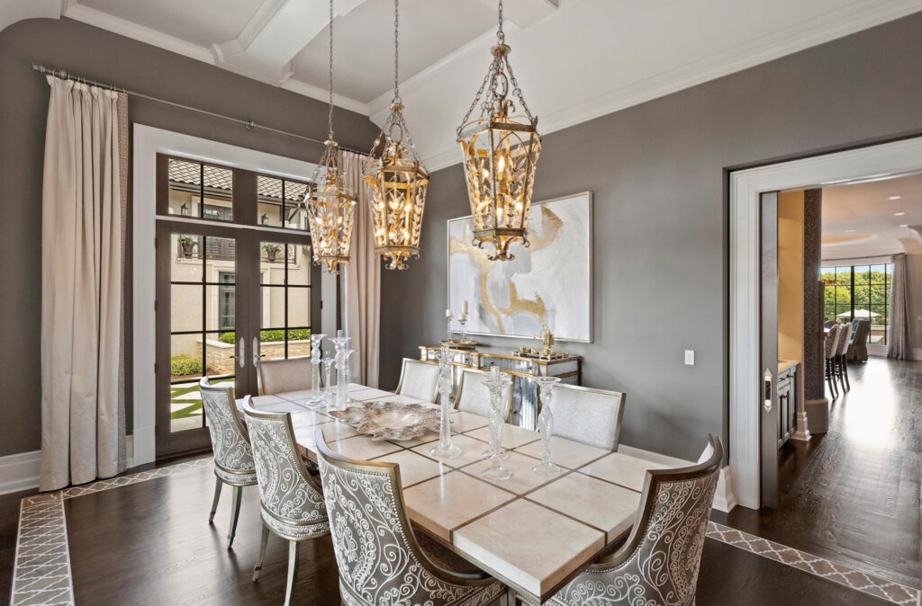 Elegant dining room with a long table, eight ornate chairs, three decorative pendant lights, large windows with curtains, modern art on the wall—a perfect example of stylish dining rooms—with views into another room and the garden outside.