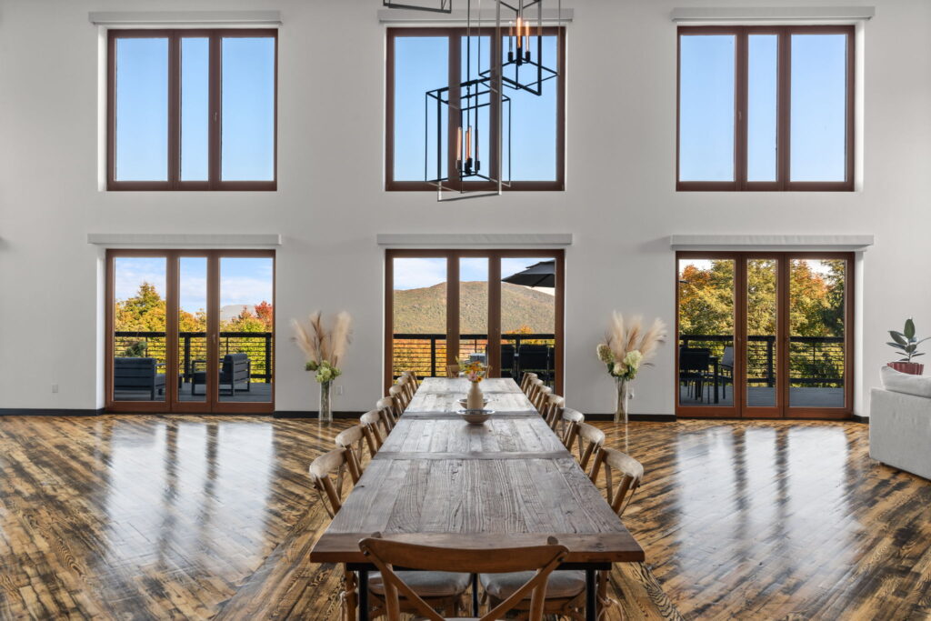 A spacious dining room with a long wooden table and chairs, large windows and glass doors filling dining rooms with natural light, offering views of a deck and green hills. The room features wooden floors and minimal decor.
