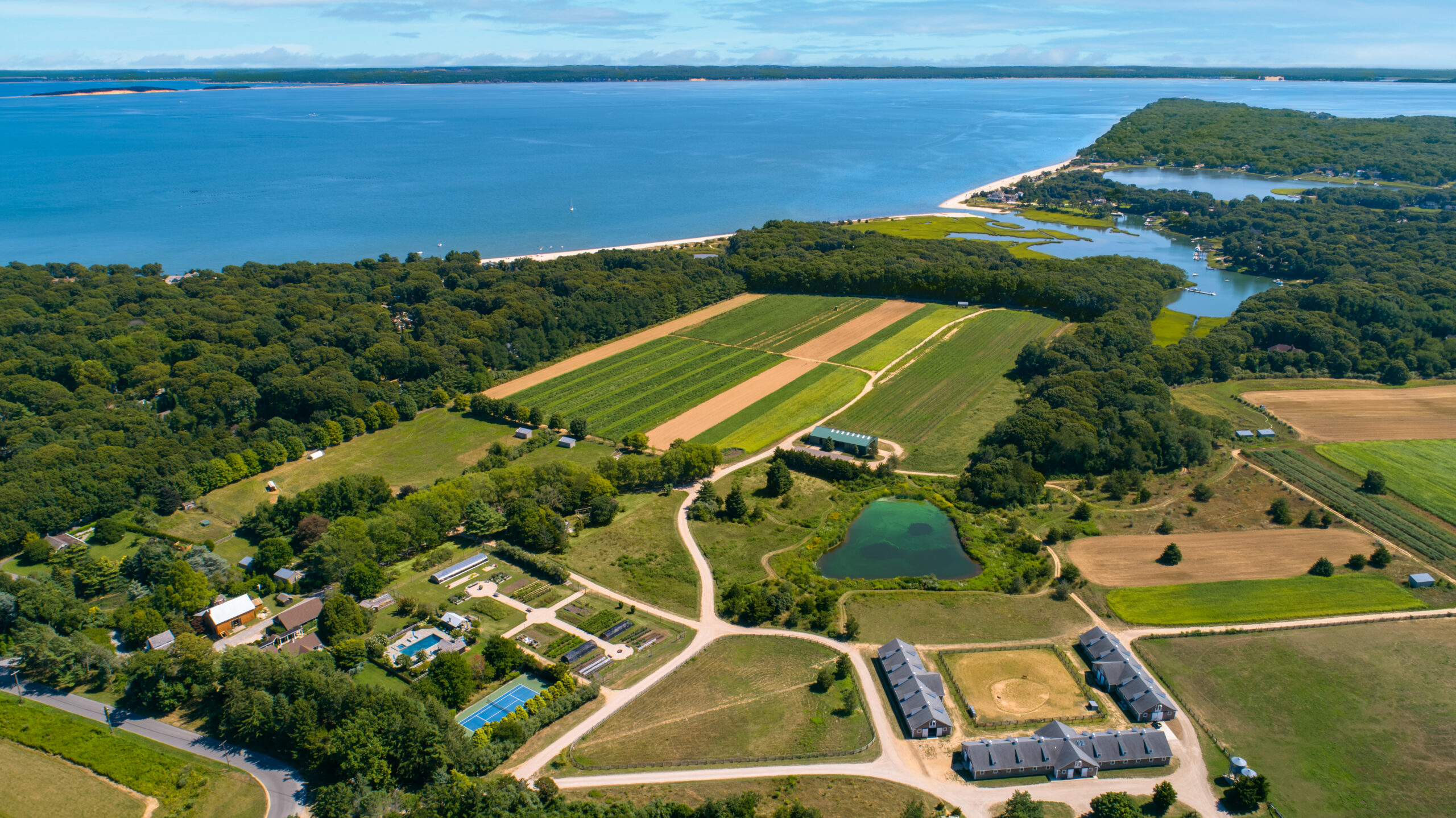 Aerial view of farmland with fields, trees, houses, and a pond near a coastline; blue water and forested land stretch into the distance under a partly cloudy sky—a captivating scene perfect for any A-list: November 2025 backdrop.