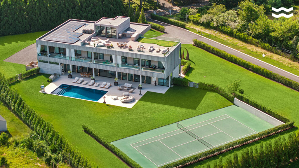 Aerial view of a modern mansion with a large lawn, swimming pool, sun loungers, rooftop seating, and a tennis court.