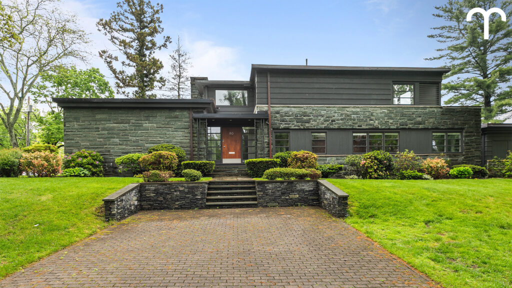 A modern two-story house with dark stone and wood exterior, large windows, a brown front door, and steps leading to the entrance—perfect for embracing new beginnings during Sagittarius season, surrounded by neatly trimmed shrubs on a green lawn.