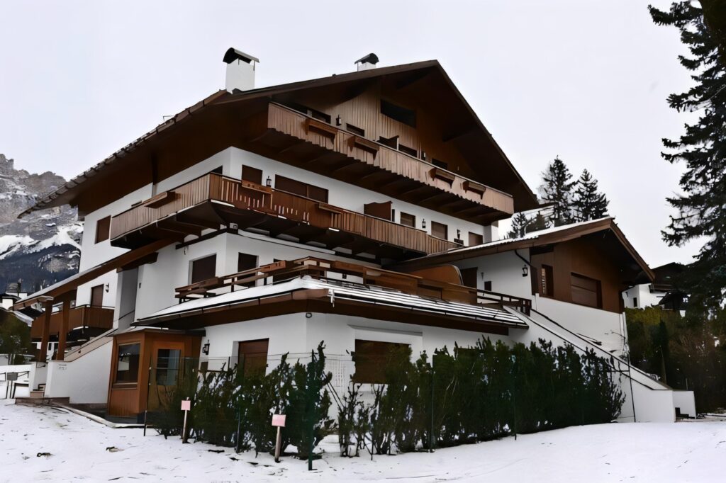 A large, alpine-style chalet with wooden balconies and a snow-covered roof stands surrounded by snow and pine trees—a striking contrast to the vibrant hues of Caribbean houses—set against a mountainous winter landscape.