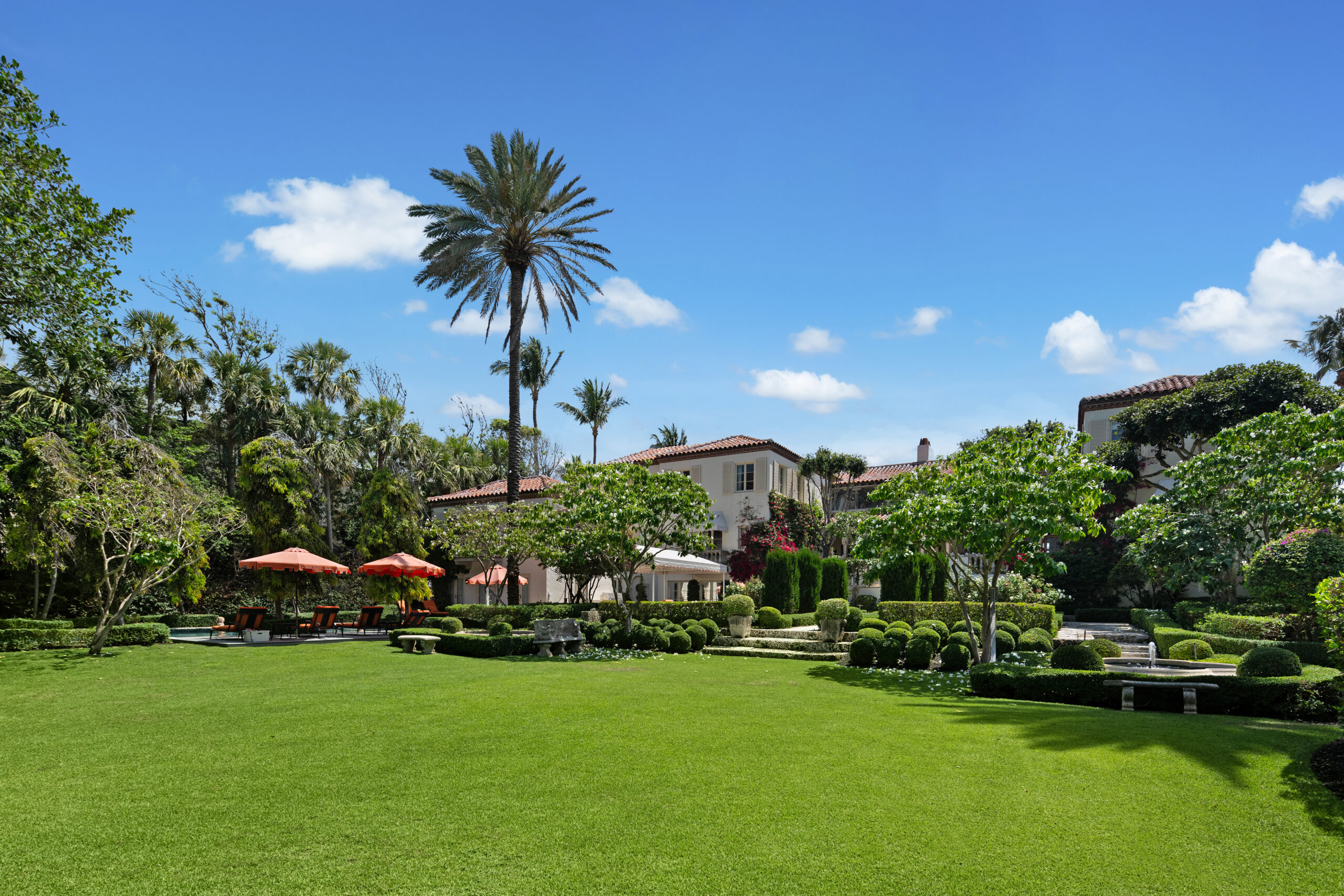 A large, elegant house with red-tiled roofs is surrounded by lush, manicured gardens and palm trees under a clear blue sky—perfect for an A-list December 2025 gathering. Red umbrellas shade lounge chairs on the patio to the left.