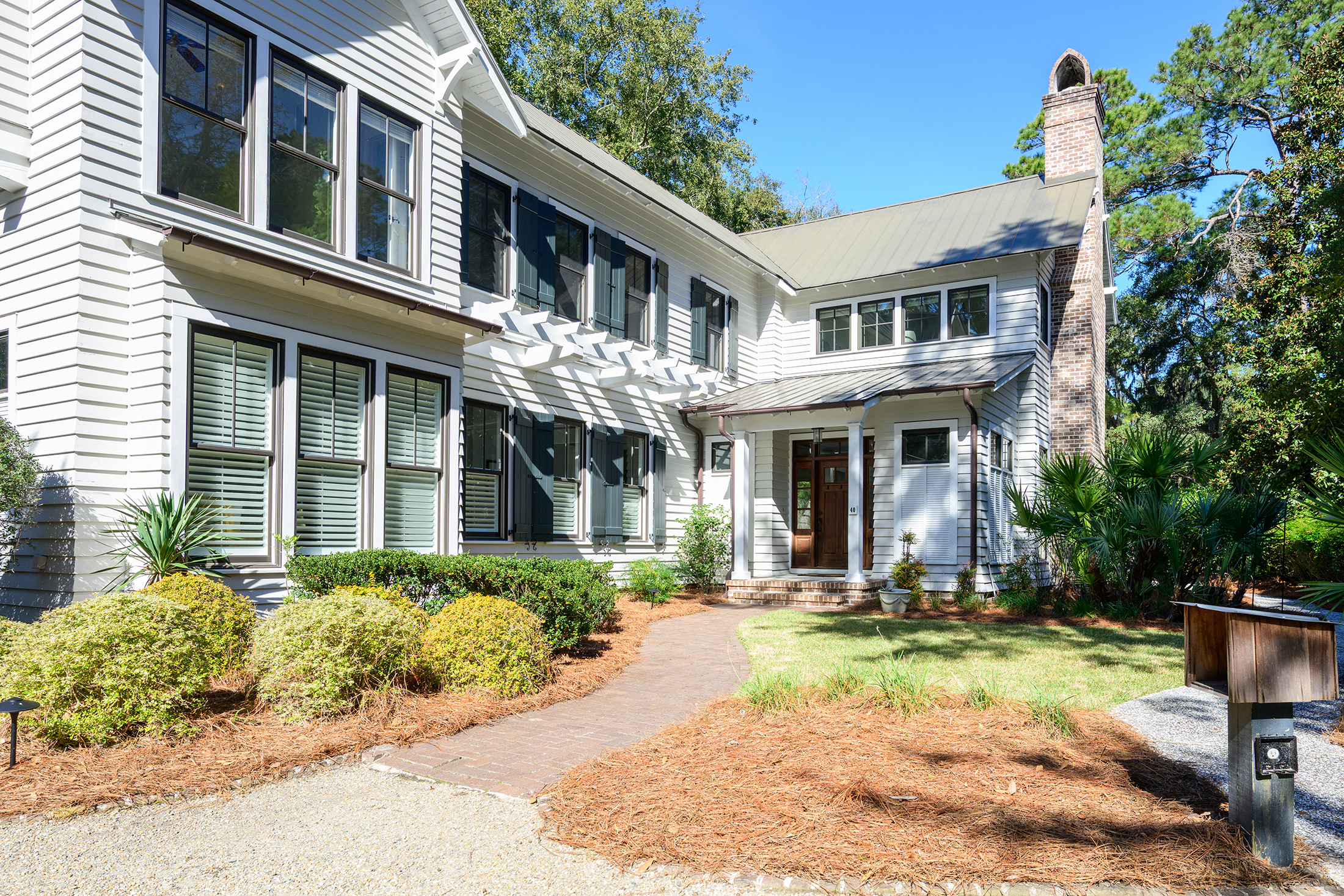 A large two-story white house with green shutters, many windows, and a brick walkway leading to the front door, surrounded by shrubs and mulch in a landscaped yard—an A-list property available November 2025.