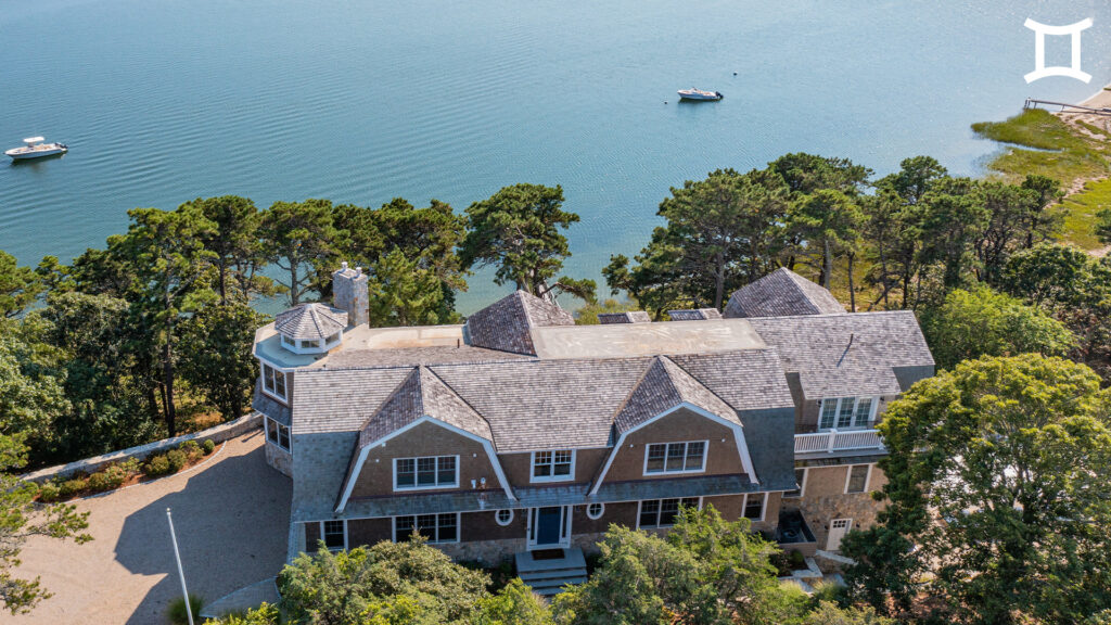 A large, shingle-style house surrounded by trees sits on the shore of a calm body of water, with boats anchored nearby. The peaceful, sunny scene feels vibrant and full of possibility—perfect for reflecting during Sagittarius season.