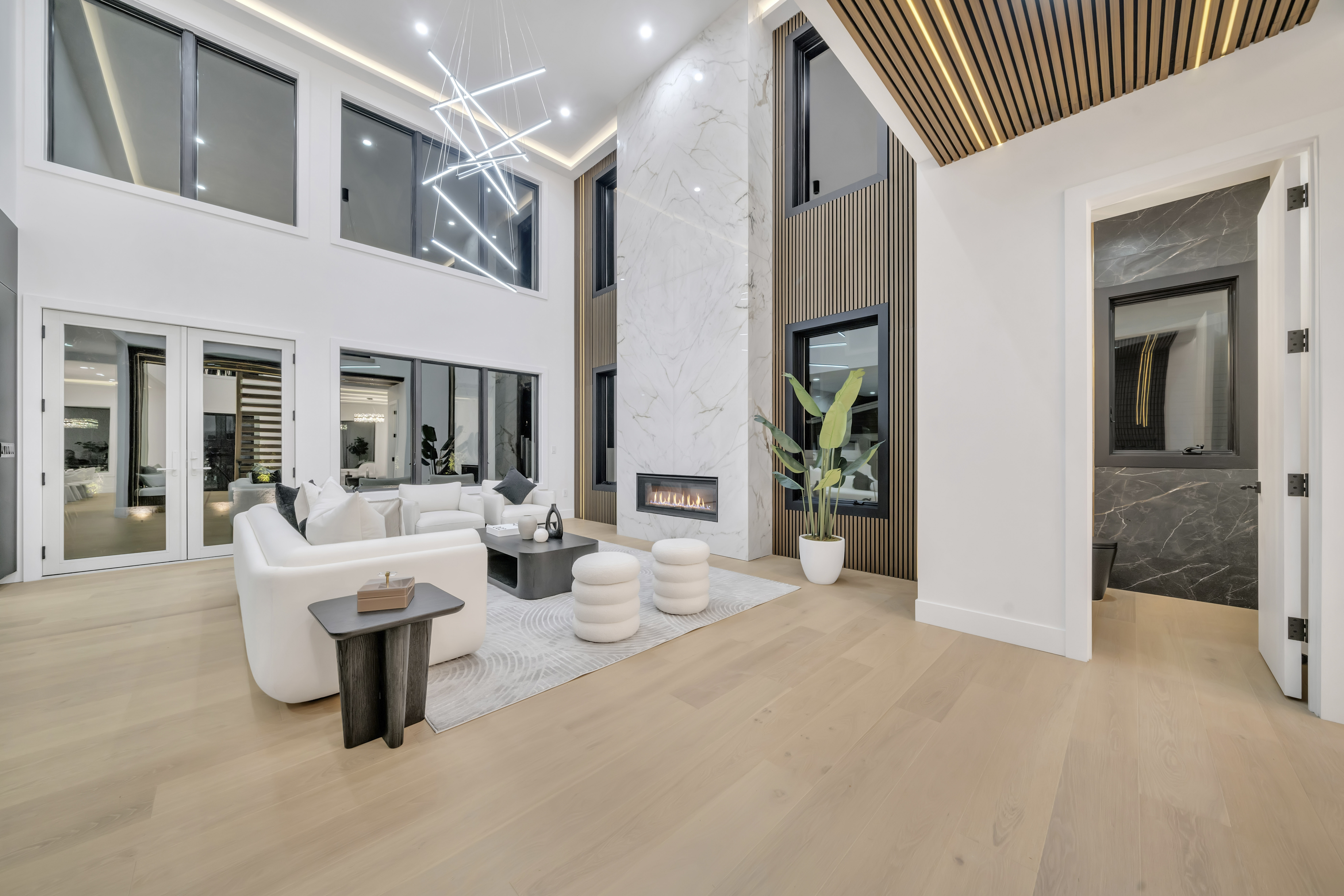 Modern living room with high ceilings, large windows, white walls, light wood floor, white sofas, round ottomans—a sleek marble fireplace and contemporary lighting create an a-list vibe perfect for December 2025 gatherings.