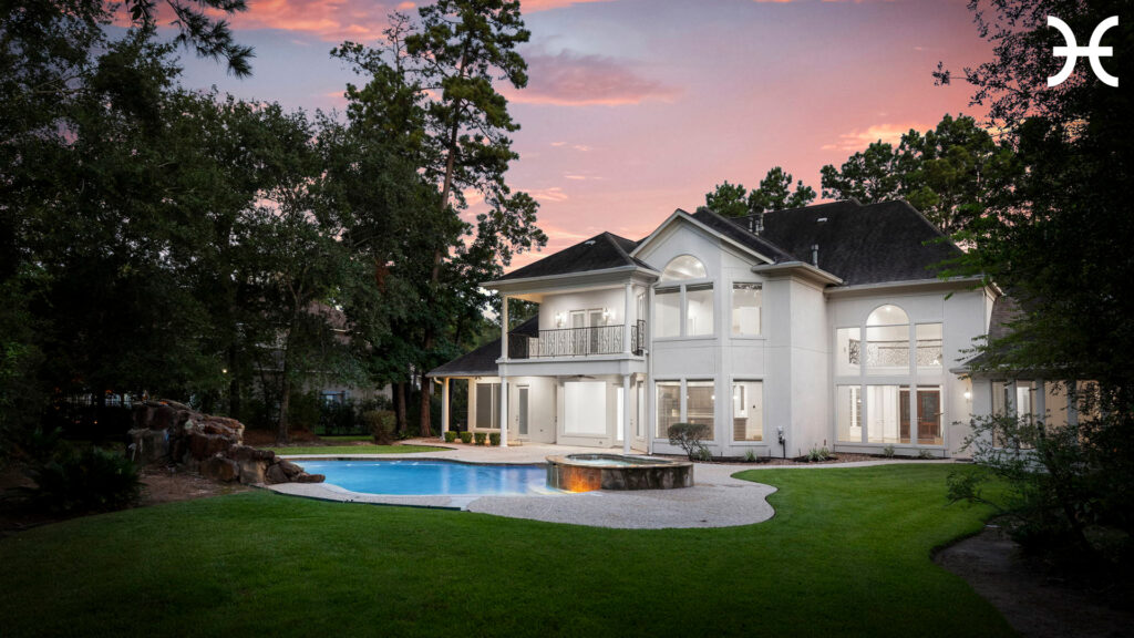 A large modern white house with many windows and balconies, surrounded by trees and a lush green lawn, features a swimming pool and spa in the backyard under a colorful sunset sky.