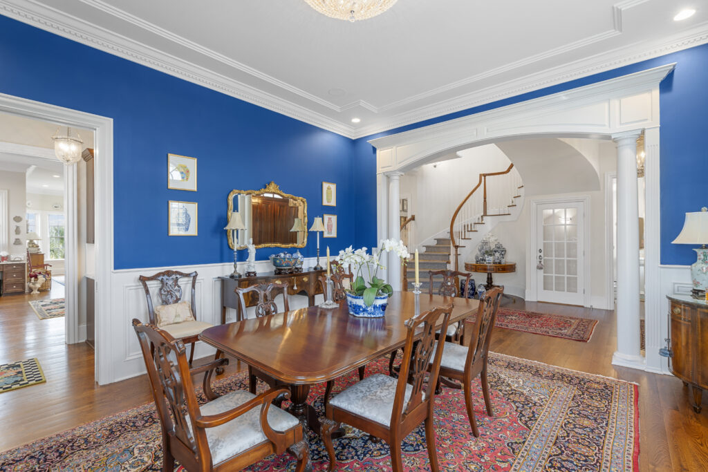 Elegant dining room with vibrant blue walls, white wainscoting, a wooden dining table for eight, decorative rugs, and classic furniture. An arched doorway reveals a staircase and entryway adorned with touches of fall foliage.