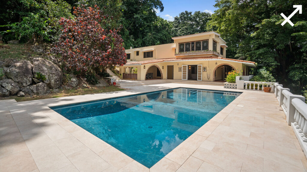 A two-story beige house with arched patios and balconies sits surrounded by trees, next to a clear blue swimming pool on a tiled terrace—perfect for relaxing under the partly cloudy sky during Sagittarius season.