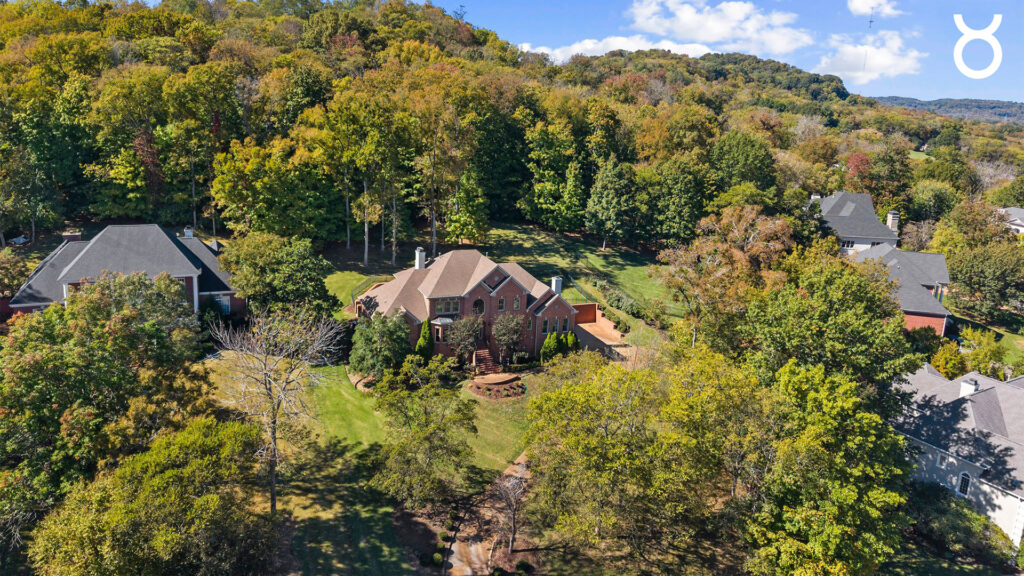 A large brick house with a spacious yard surrounded by trees, set in a lush, green, hilly neighborhood. Perfect for enjoying the fresh air during Sagittarius season, with wooded hills and other homes visible under a partly cloudy sky.