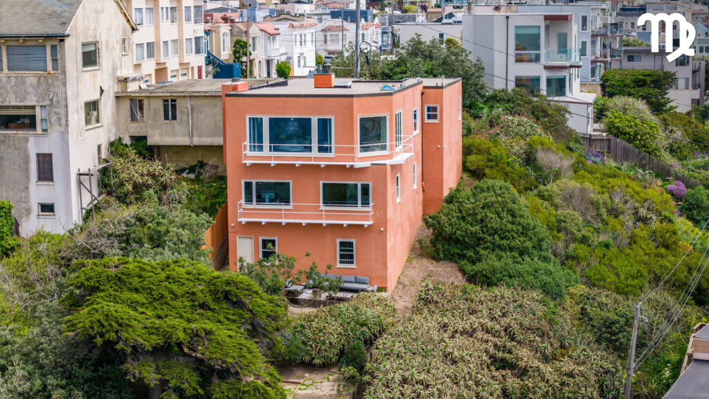 A peach-colored, three-story house with white trim sits on a green, overgrown hillside surrounded by other homes in a densely built urban neighborhood. The Virgo zodiac symbol and a nod to astrological houses appear in the top right corner.