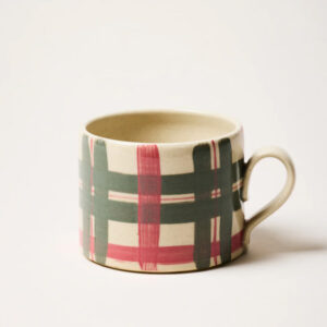 A ceramic mug with a handle, featuring a plaid pattern of wide green and red brushstrokes on a cream-colored background, set against a plain white backdrop—perfect for enjoying coffee while browsing your favorite cookbooks.