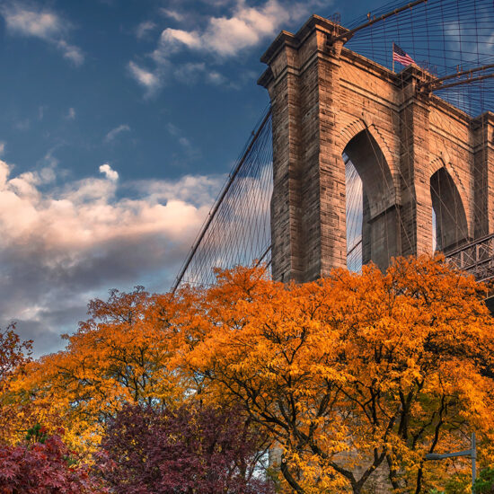 The Brooklyn Bridge rises above vibrant autumn trees with orange and yellow leaves, set against a partly cloudy blue sky in the early evening—a picturesque scene reflecting the allure of Manhattan luxury sales.