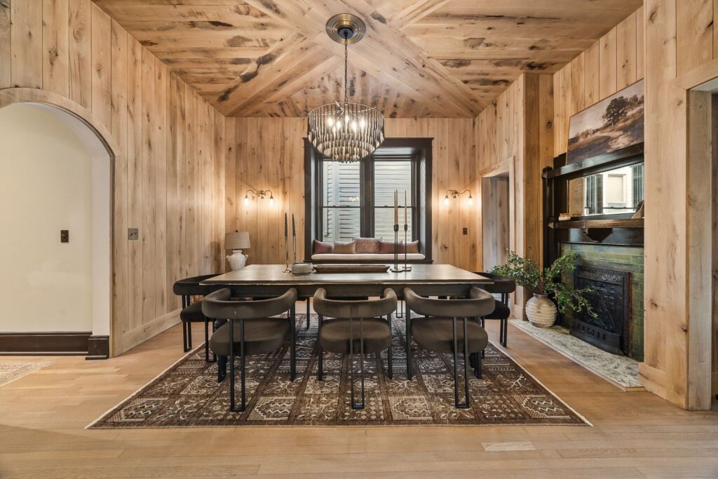 A cozy dining room with wood-paneled walls and ceiling, a long black dining table with eight chairs, a patterned rug, modern chandelier, wall sconces, and a decorative fireplace—an inviting space that defines stylish dining rooms.