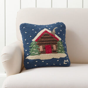 A square decorative pillow featuring a cozy log cabin with a red door, surrounded by green trees and snow, set against a blue background with white snowflakes—perfect for curling up with your favorite cookbooks on a light-colored couch.