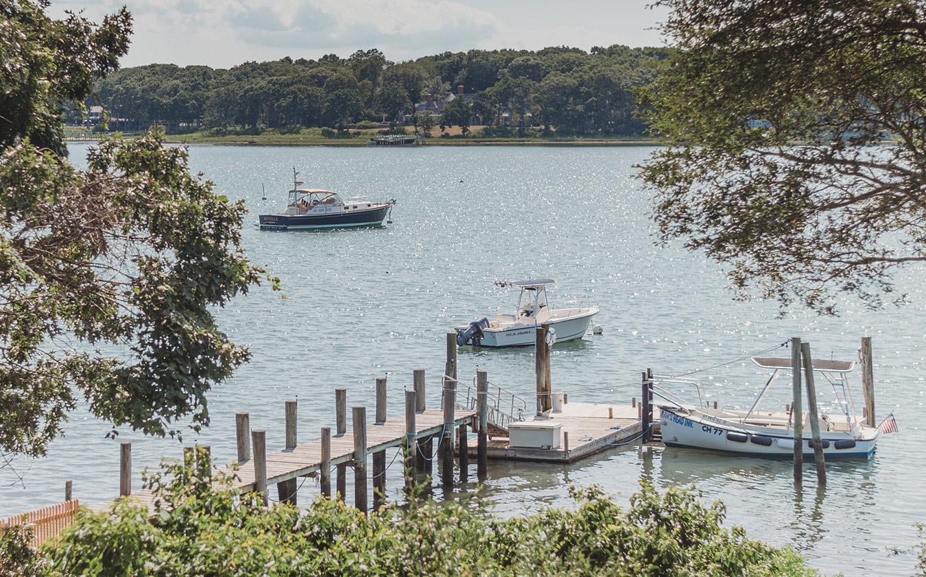 A small wooden dock extends into a calm lake with three boats, two near the dock and one farther out. Trees frame the view and a wooded shoreline is visible in the distance under partly cloudy skies.