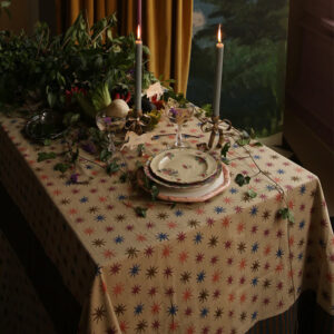 A dimly lit table set for one with a star-patterned tablecloth, a floral plate, silverware, cookbooks stacked nearby, two glasses, two burning candles, and greenery draped across the tabletop.