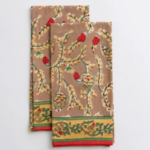 Two folded brown cloth napkins with a floral and leaf pattern in red, green, and yellow, and a decorative yellow and green border at one end, placed on a white surface beside vintage cookbooks.