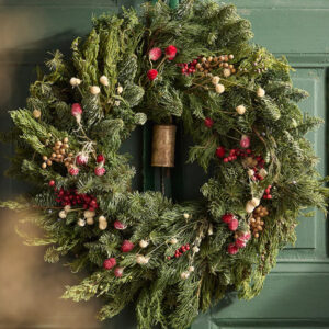 A festive green wreath, reminiscent of classic holiday cookbooks, decorated with red and white berries and small pinecones hangs on a dark green door.