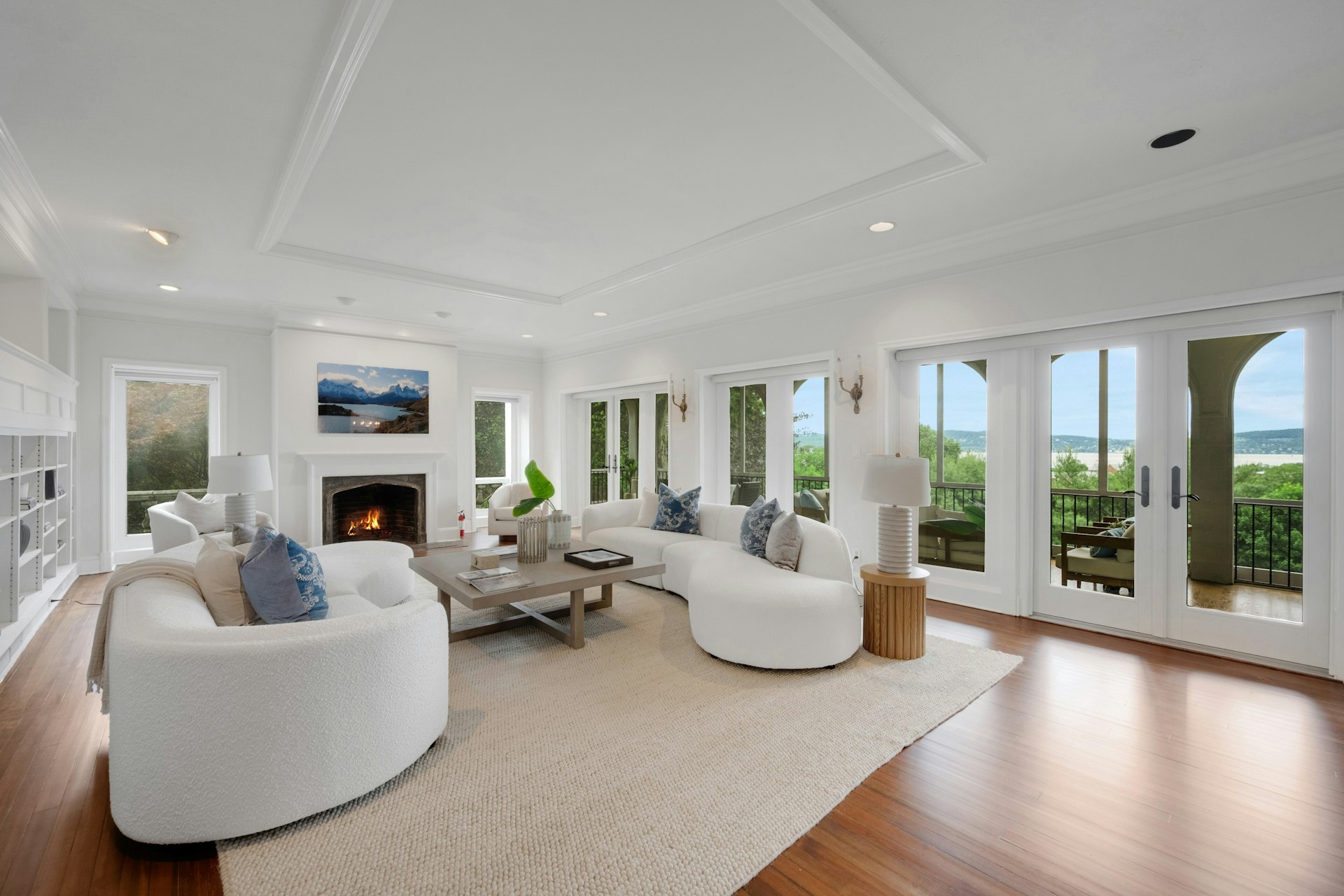 028-480012-MTZ_5908_14150208 Spacious, bright living room with white walls and furniture, a fireplace, wooden floors, and large windows—a perfect A-list: January 2026 retreat—plus glass doors opening to a scenic balcony overlooking greenery and water. A beige rug anchors the modern seating area.