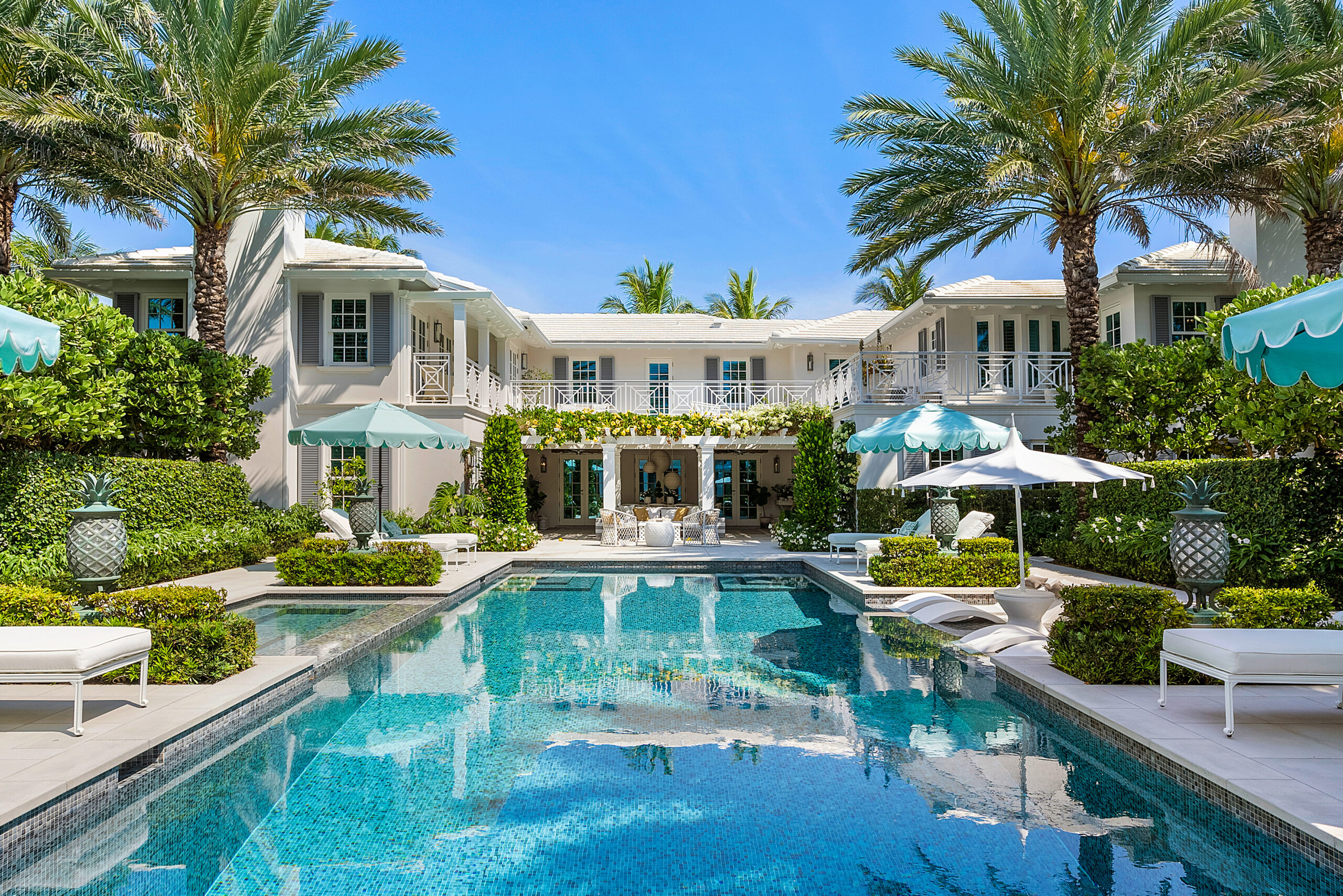 050-880SouthOceanBlvd-PalmBeach-FL-33480-FULL A luxurious villa with white exterior, surrounded by palm trees, features a large swimming pool and stylish lounge chairs—an a-list escape for January 2026. The house boasts balconies and an open patio area under the sunny sky.