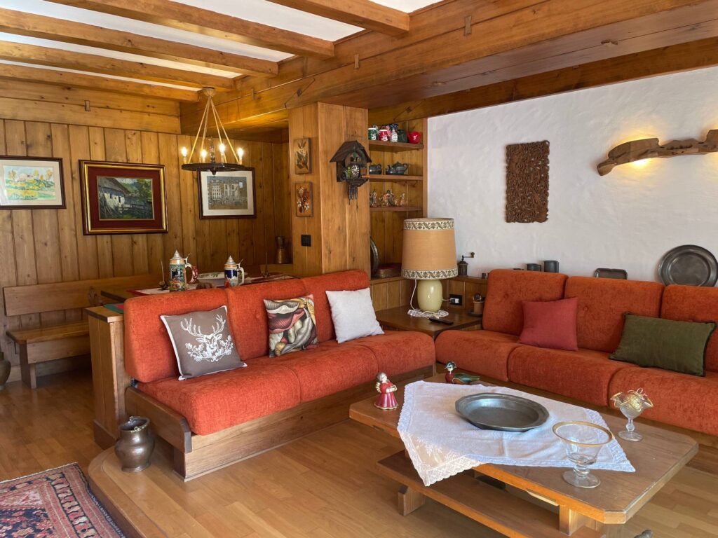 A cozy living room with wood-paneled walls and ceiling, orange sofas with decorative pillows, a coffee table set for guests, and shelves displaying figurines—evoking the vibrant charm often found in Caribbean houses. Paintings and art decorate the walls.