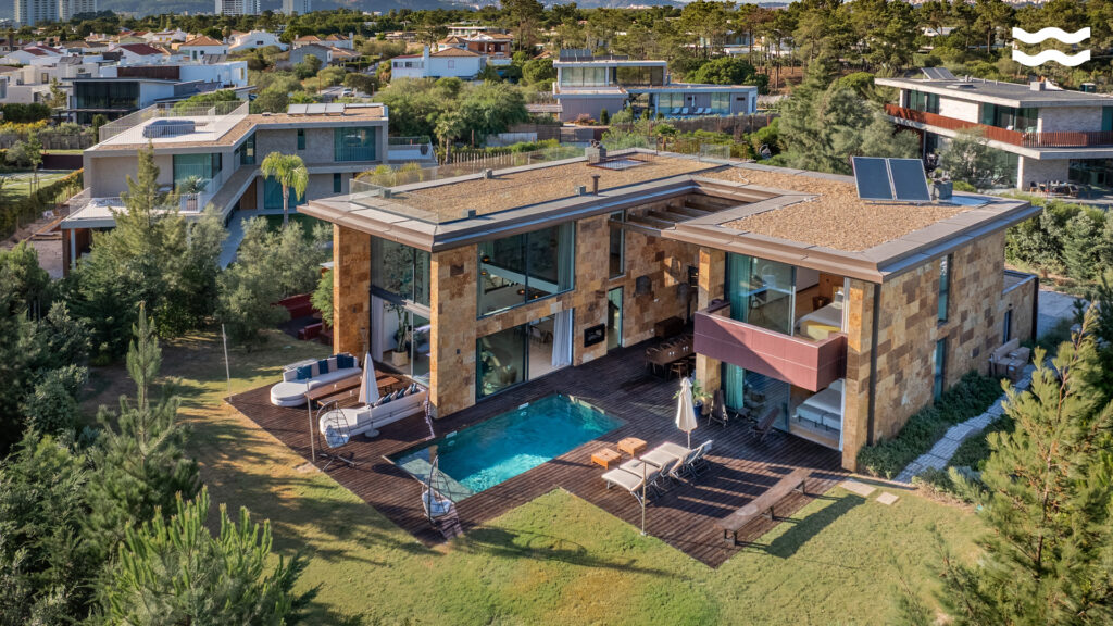 Aerial view of a modern two-story house with stone exterior, large windows, a small pool, and outdoor seating—perfect for relaxing during Capricorn season—nestled among greenery and neighboring contemporary homes.