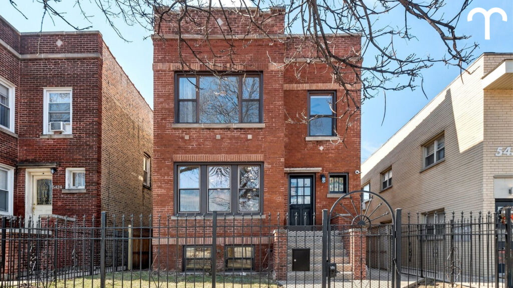 A two-story red brick house with large windows, a black metal fence, and a gated entrance stands between two neighboring buildings on a sunny Capricorn season day.