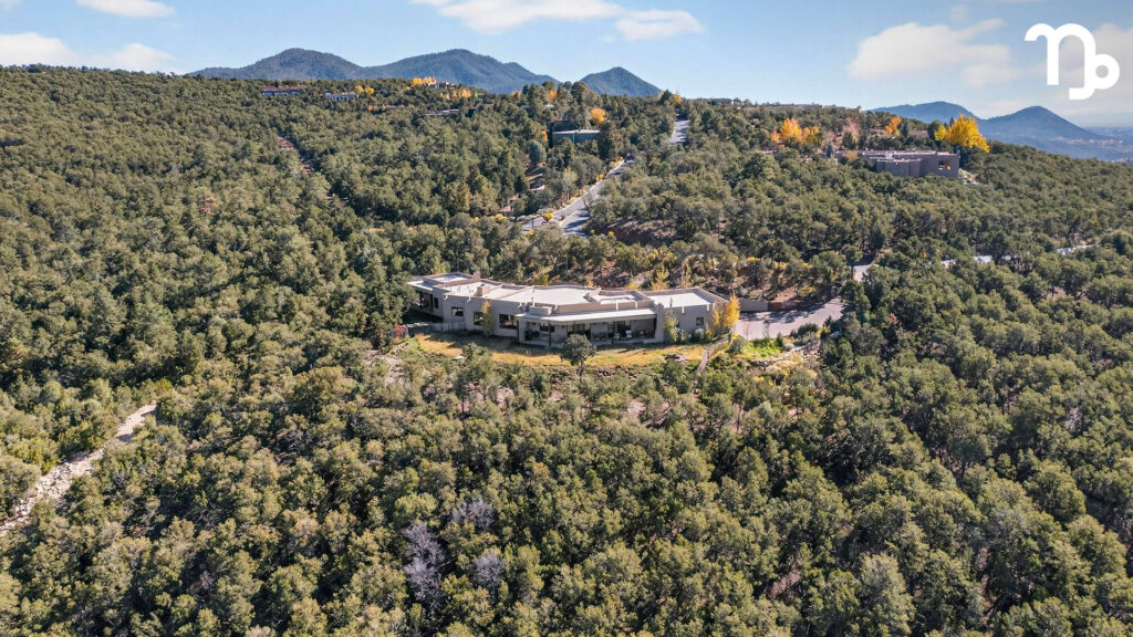 A large modern house is nestled among dense green trees on a hillside, with winding roads and more homes visible in the background. Mountain peaks rise in the distance under a clear blue sky, hinting at Capricorn season and its connection to astrological houses.