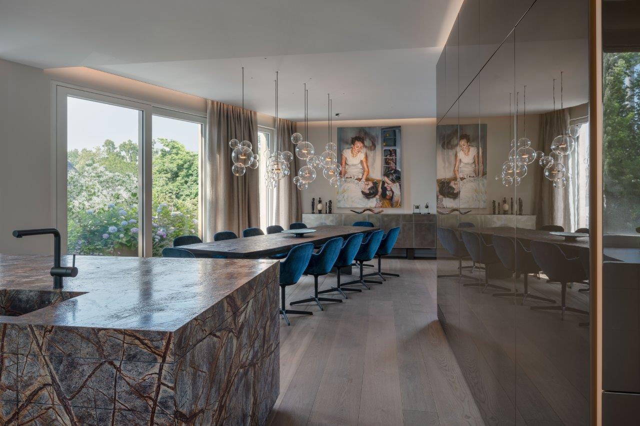 Foto 3 innen Modern dining area with a stone island, blue velvet chairs, glass pendant lights, and large windows showcasing a garden view. Minimalist decor and stunning portrait artwork evoke an a-list: january 2026 sophistication.