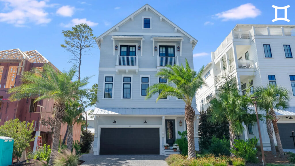 A modern three-story white house with balconies, palm trees, and a dark double garage stands tall—perfect for Capricorn season. Neighboring homes and a house under construction are visible on either side under a blue sky.