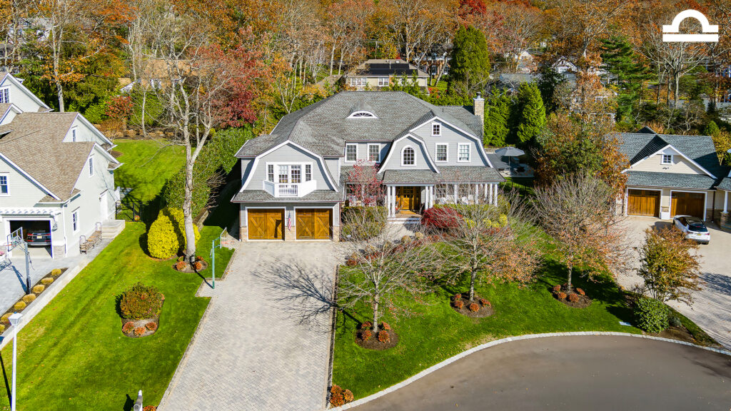 A large two-story gray house with white trim and three wooden garage doors sits on a landscaped lot, its green lawns and trees reflecting the crisp beauty of Capricorn season in a suburban neighborhood during autumn.