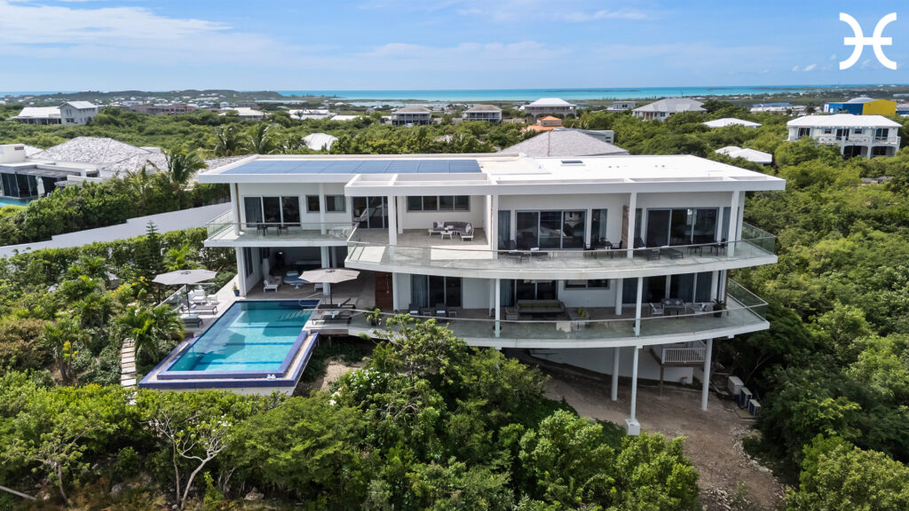 A modern, two-story villa with large glass windows, spacious balconies, and an infinity pool overlooks lush greenery and neighboring houses, creating a serene retreat perfect for reflection during Capricorn season—with the ocean visible in the background.