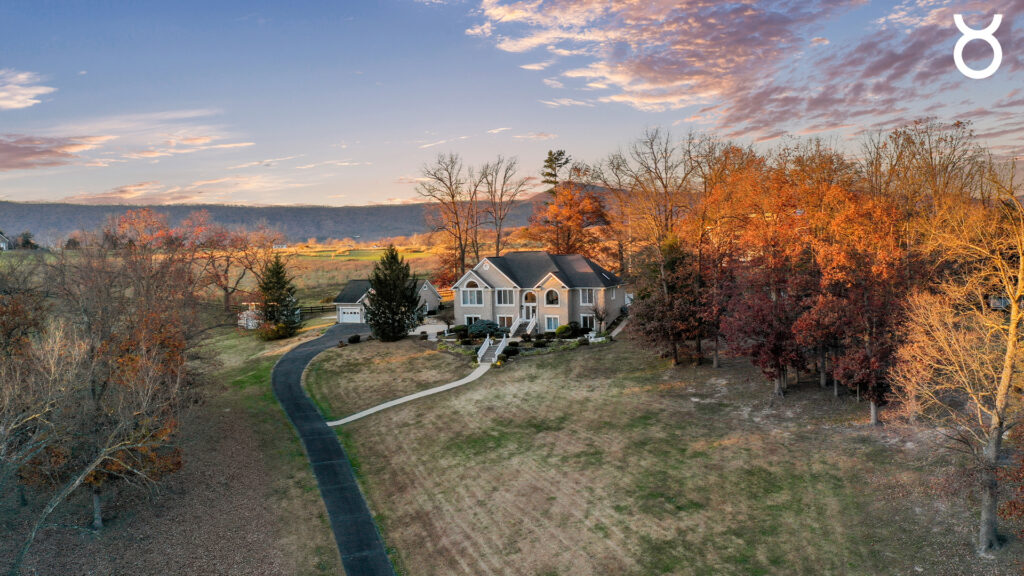 A large beige house with tall windows sits on a spacious lot with autumn trees. During Capricorn season, a curving driveway leads to the house, set against distant hills under a partly cloudy sky at sunset.