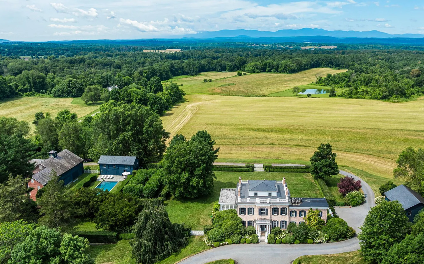 A large country estate with a main house, swimming pool, and outbuildings surrounded by trees, gardens, and open fields, with hills and mountains visible in the distance under a blue sky.