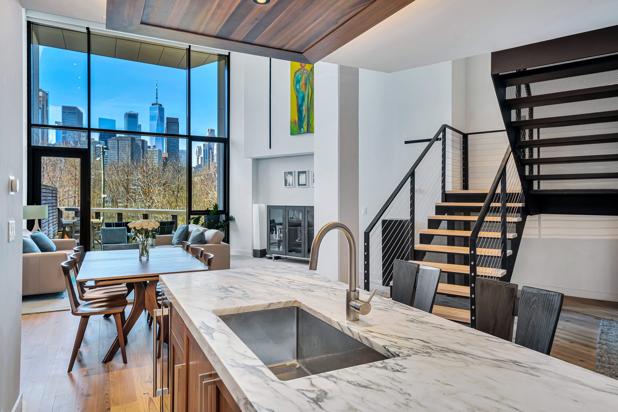 Photos of 90 Furman St Apt 220 for broker Abigail Perlman of Douglas Elliman Modern open-plan living space with a marble kitchen island, dining table, and lounge area, large floor-to-ceiling windows showing a city skyline, and a staircase leading to an upper floor—an A-list look for January 2026.
