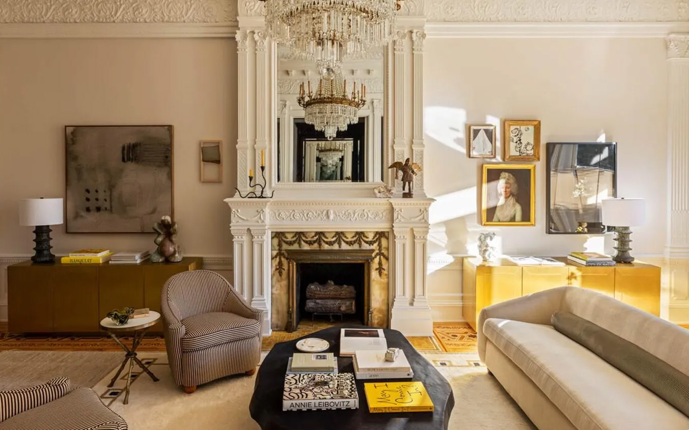 Elegant living room with ornate white fireplace, crystal chandelier, modern furniture, books on a central coffee table, artwork and portraits on cream-colored walls, and sunlight streaming through large windows.
