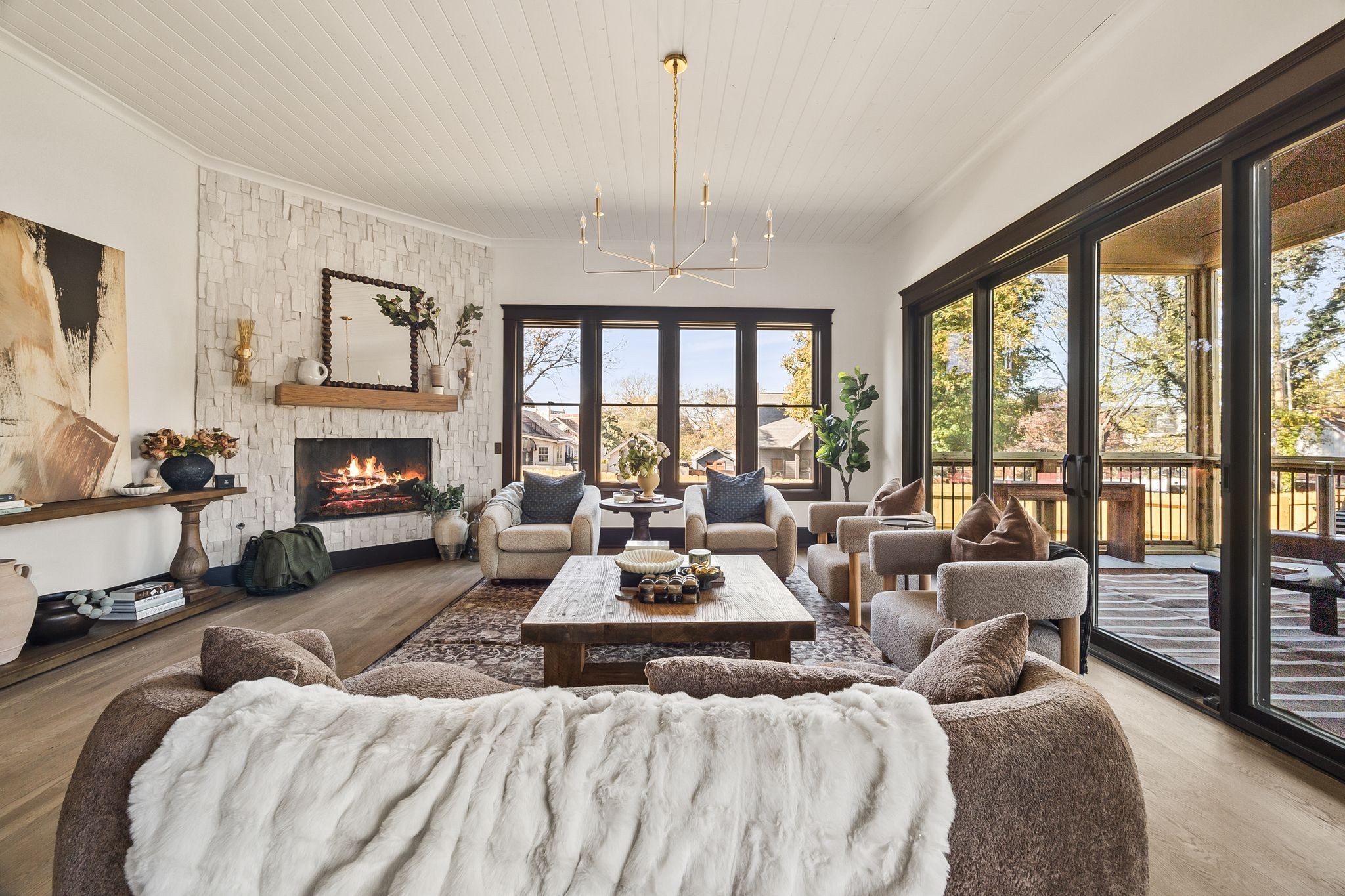 photo_online_027 A modern living room with neutral tones, cozy armchairs, a sofa with a fur throw, and a coffee table sits by the fireplace. Large windows open to a deck with trees visible outside—perfect for relaxing or welcoming your A-list guests this January 2026.