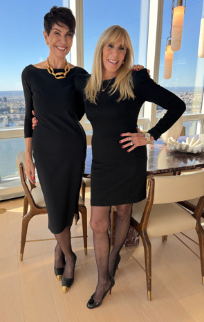 Two women stand smiling in a bright room with large windows. Both wear black dresses and black heels. The woman on the left has short dark hair and gold jewelry; the woman on the right has long blonde hair.
