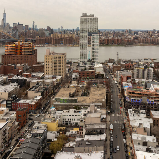 Aerial view of Williamsburg, Brooklyn, with mid-rise buildings and brooklyn condos lining the streets. The East River glimmers in the background, while Manhattan’s skyline and the Empire State Building rise under a cloudy sky.