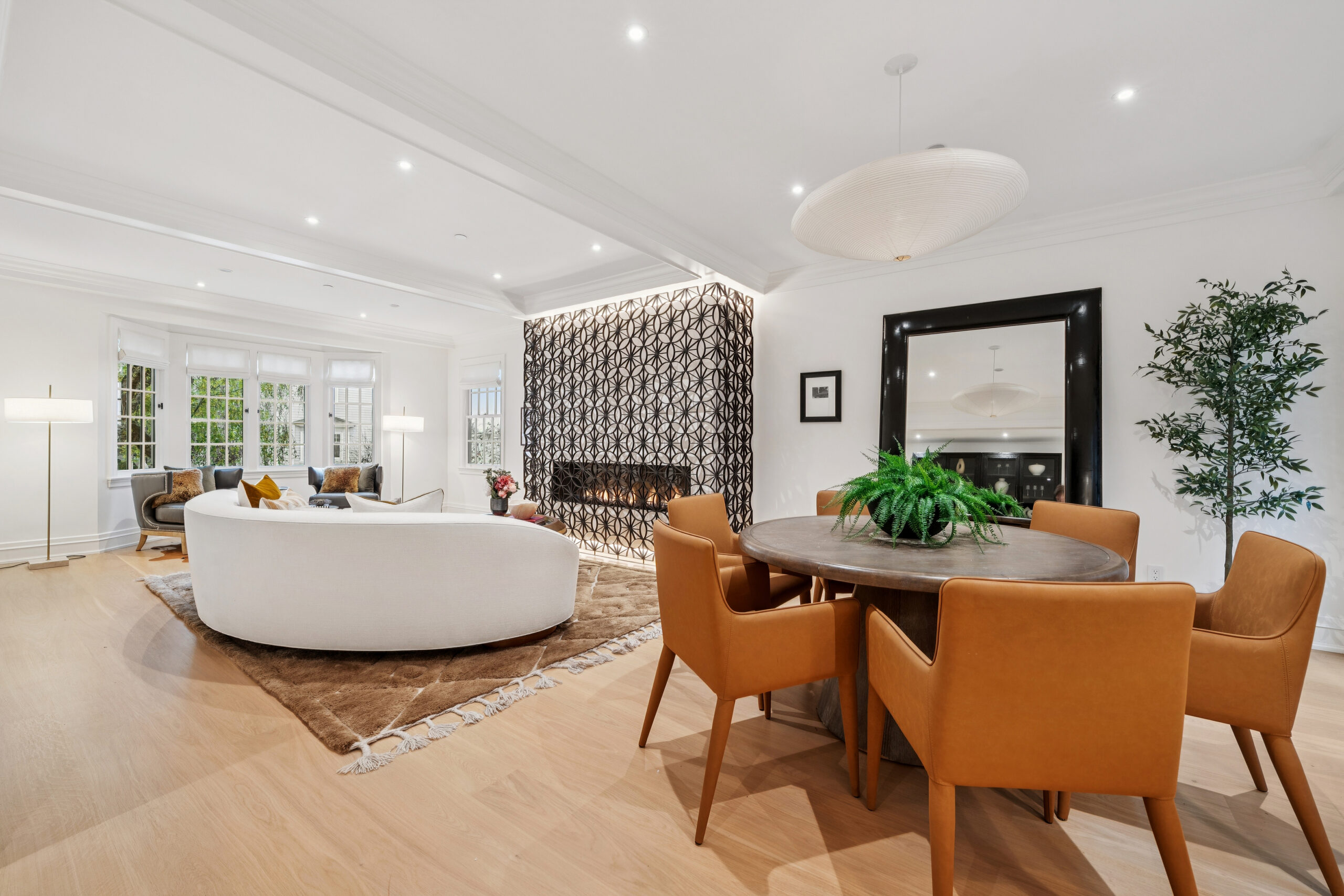 A modern open living and dining area with a white curved sofa, round wooden table with tan chairs, patterned accent wall with fireplace, large mirror, potted plant, and hardwood floors—a-list: February 2026 design inspiration.