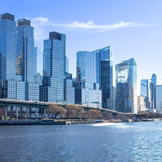 A city skyline with tall, modern glass buildings lines the waterfront under a clear blue sky. In the foreground, a river flows past a highway and scattered trees, evoking the vibrant energy of a Brooklyn condo setting.