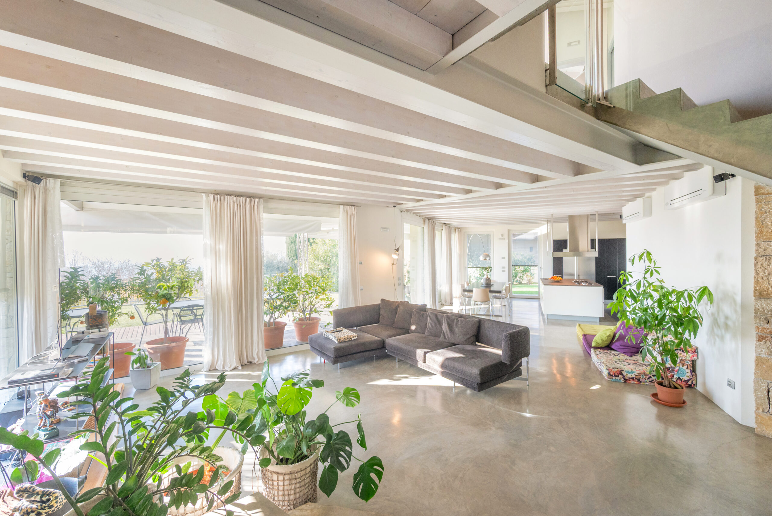 Spacious, sunlit living room with large windows, gray sectional sofa, indoor plants, and polished concrete floors—an A-list February 2026 retreat. Open views to the kitchen and dining area plus stairs leading to a cozy reading nook with cushions.