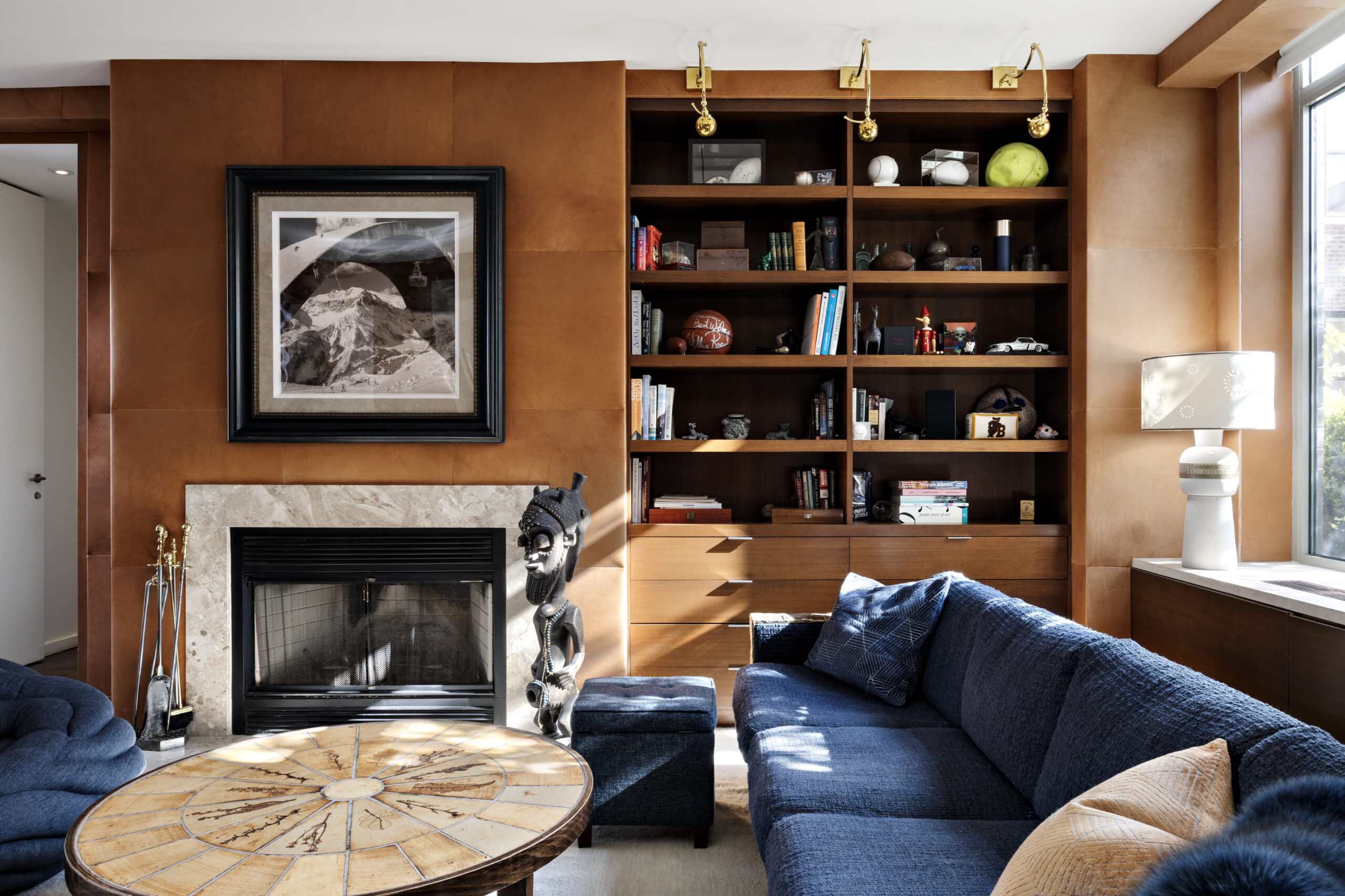 A cozy living room with a blue sectional sofa, a round wooden coffee table, and built-in bookshelves filled with books and decor. Sunlight streams in through a large window, highlighting the fireplace with a framed photo above it.