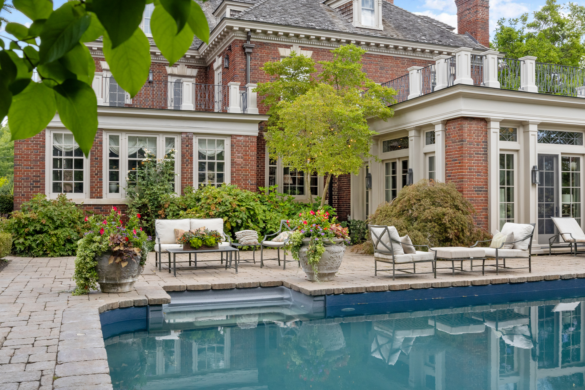 A brick house with large windows overlooks a patio with outdoor seating and potted plants, surrounded by lush greenery beside a clear blue swimming pool—perfect for relaxing A-list style in February 2026.