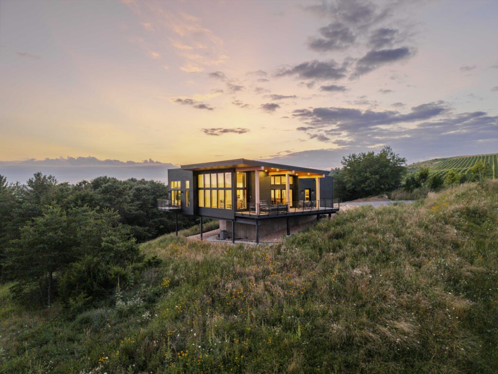 A modern house with large windows sits on a grassy hill at sunset, surrounded by trees and wildflowers, under a soft, colorful sky inspired by Aquarius season and distant fields in the background.