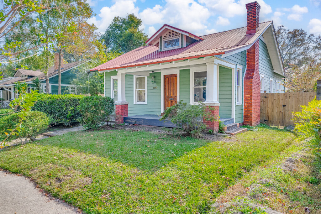 A small green Craftsman-style house with white trim, a wooden front door, red brick chimney, and porch sits surrounded by a neatly trimmed lawn and bushes, radiating cozy charm—perfect for reflecting on astrological houses during Aquarius season.