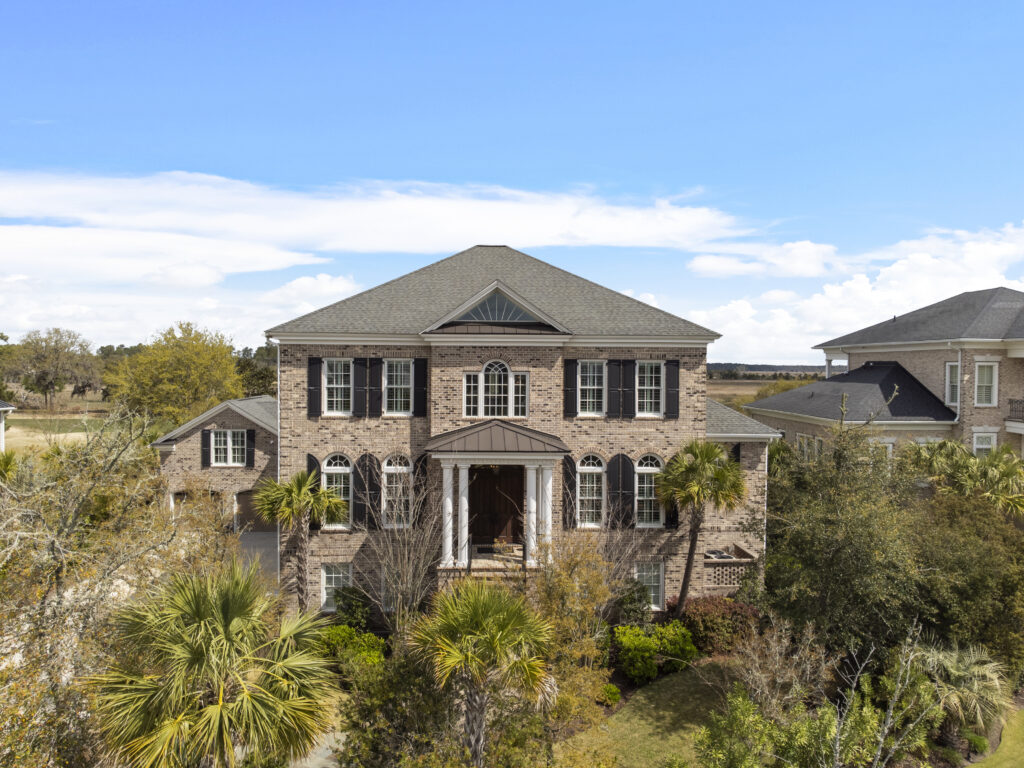 A large two-story brick house with black shutters, arched windows, and a front porch, surrounded by palm trees and greenery under a blue sky—perfect for relaxing during Aquarius season.