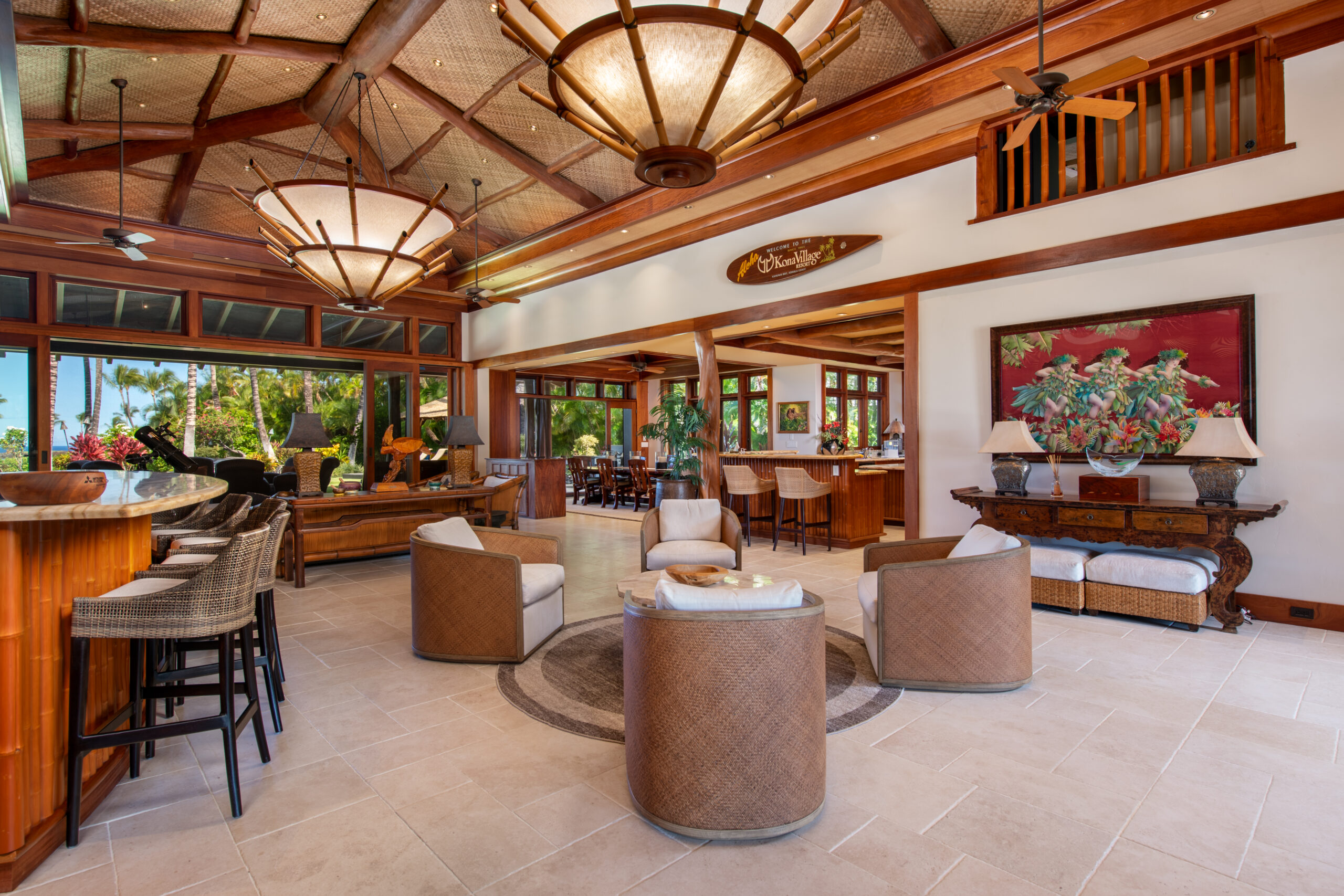 Spacious, tropical-style living room with woven chairs, a round rug, and wooden bar stools. Large windows, ceiling fans, and tropical decor create a bright space—perfect for an a-list: February 2026 gathering amid art and lush plants.